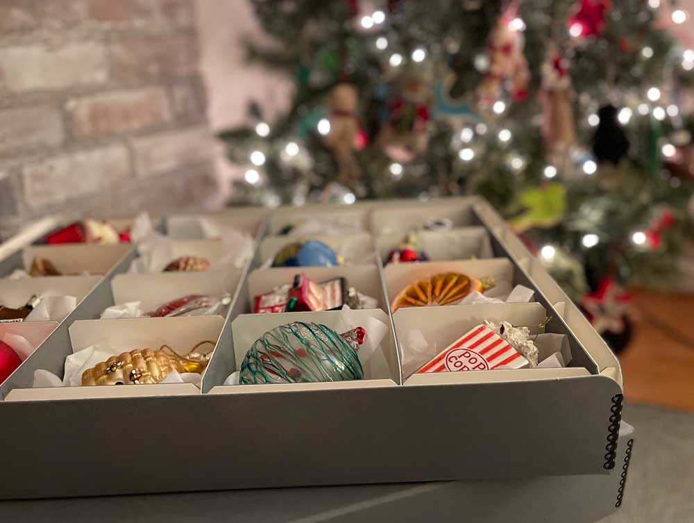 Archival Methods gray divided storage box with glass Christmas ornaments in front of a decorated Christmas tree. 