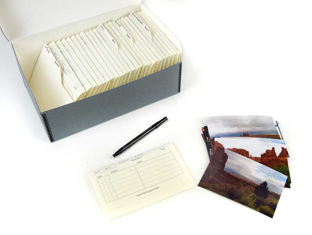Metal edge box holding acid-free archival photo envelopes and index cards with a stack of photos and pen next to it.